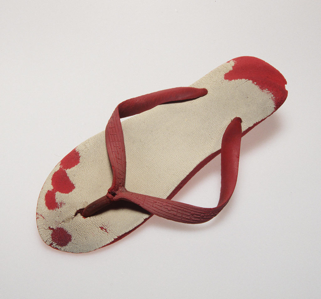 A single worn rubber thong from a pair with red soles and straps and white upper soles. The straps are moulded with musical notation motifs. The thong is so well worn to the point that the imprint of the heel and individual toes are worn into the red rubber. 